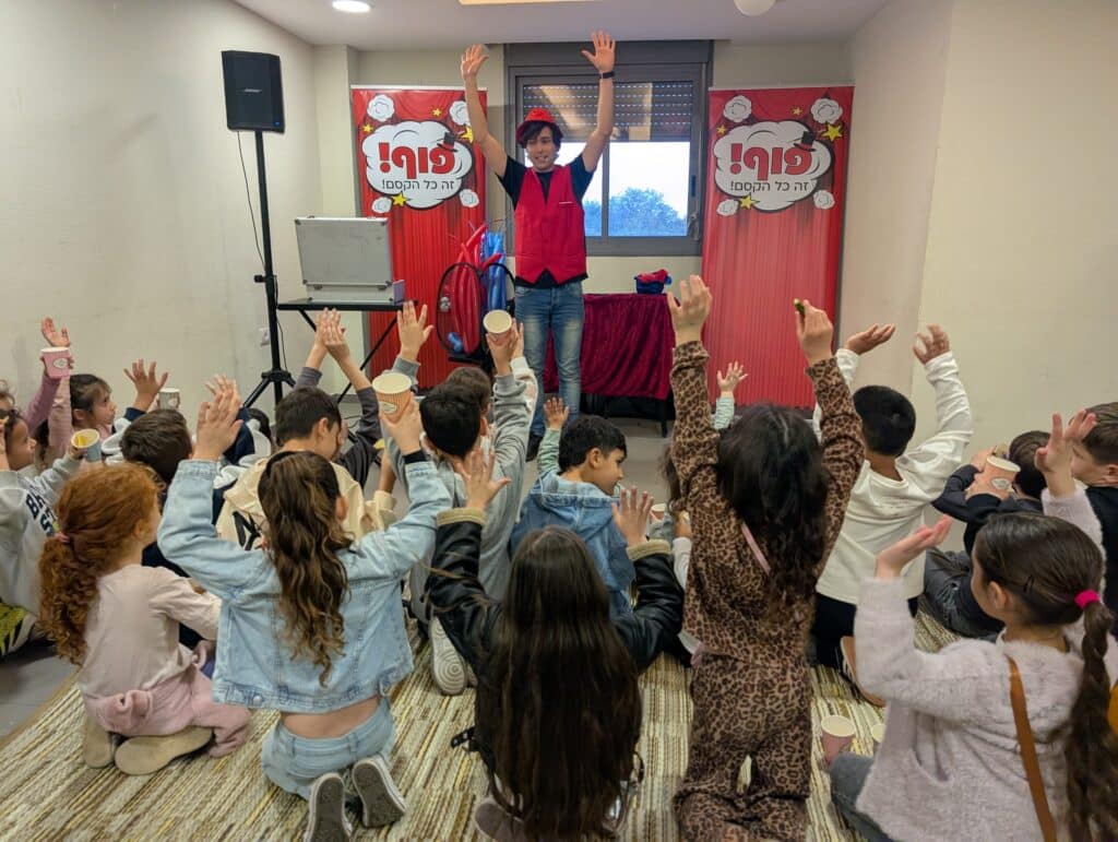 קוסם ילדים עומד עם ידיים מורמות מול קבוצת ילדים שיושבים על הרצפה ומשתתפים בפעילות עם ידיים באוויר.